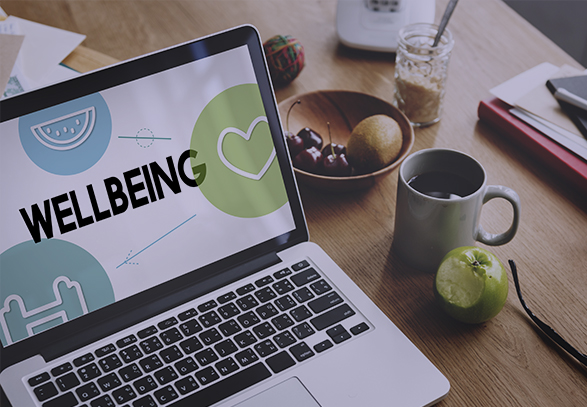 laptop open on a desk with 'wellbeing' written on the screen with some fruit in a bowl on the table next to it